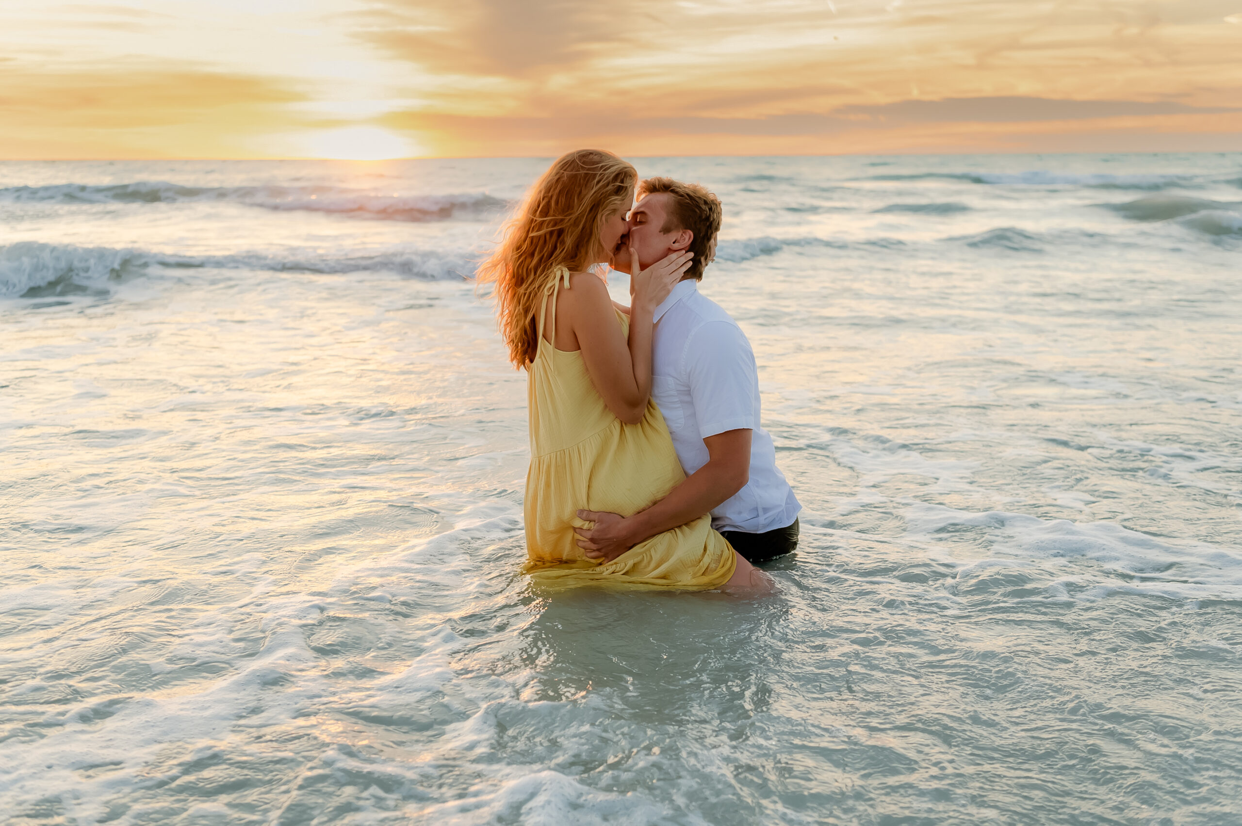 adventure beach engagement session with sunset _ couple kissing and hugging in water with water covering their legs and clothes to their knees _ woman in yellow sun dress and man in white shirt and jeans _ style by caitlin anne