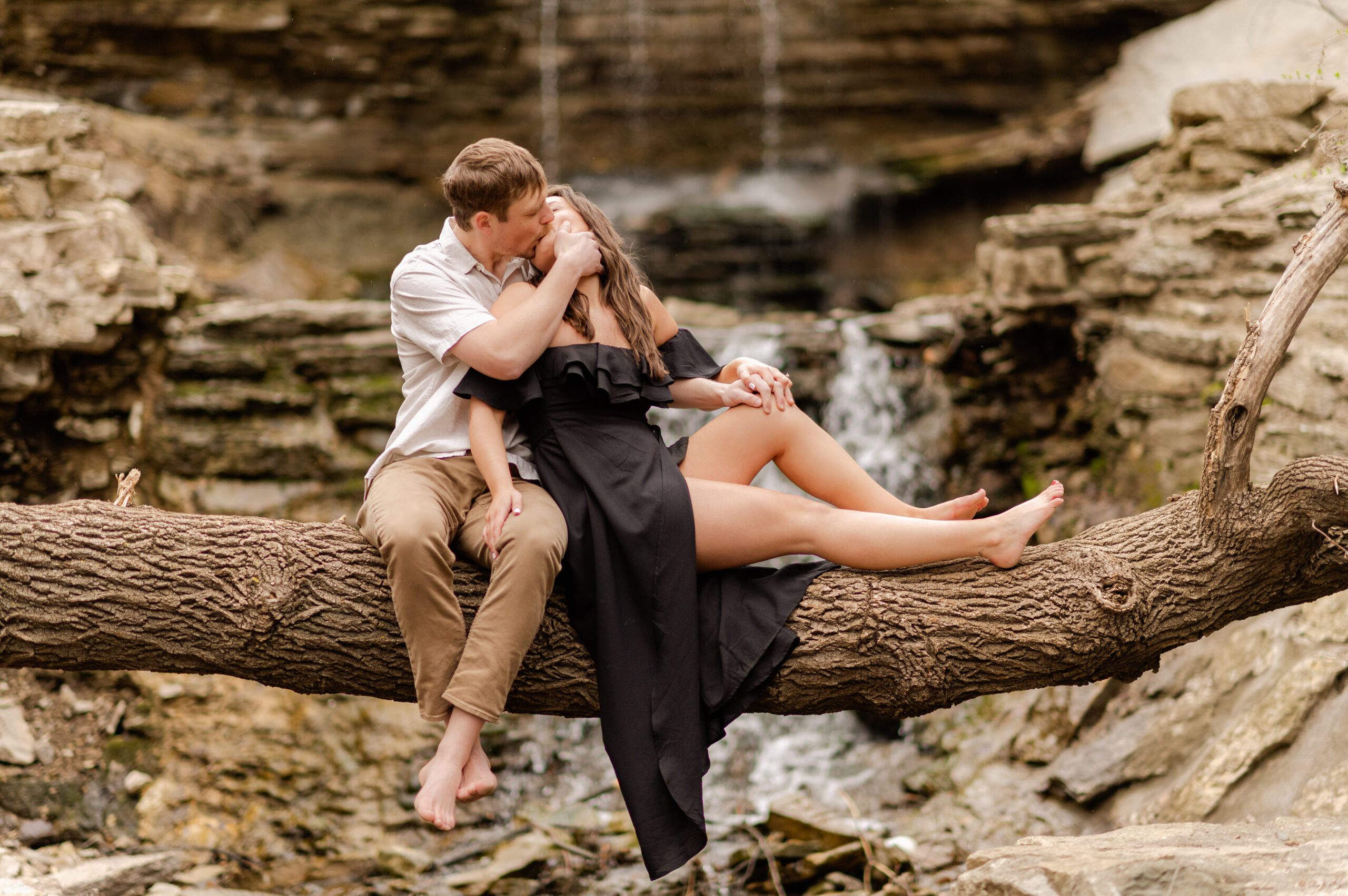 adventure engagement session at creek with waterfall and tree bridge couple sitting on tree bridge in front of waterfall kissing man in khakis and white shirt and woman in black off shoulder dress styled by caitlin anne